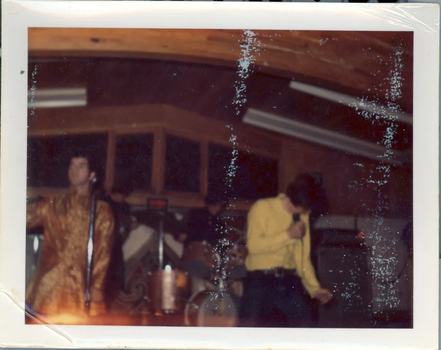 Color print with silver emulsion damage: on stage under a wood-paneled ceiling, a singer in a gold brocade jacket at the microphone at left while another performer in yellow dances at right, drums behind