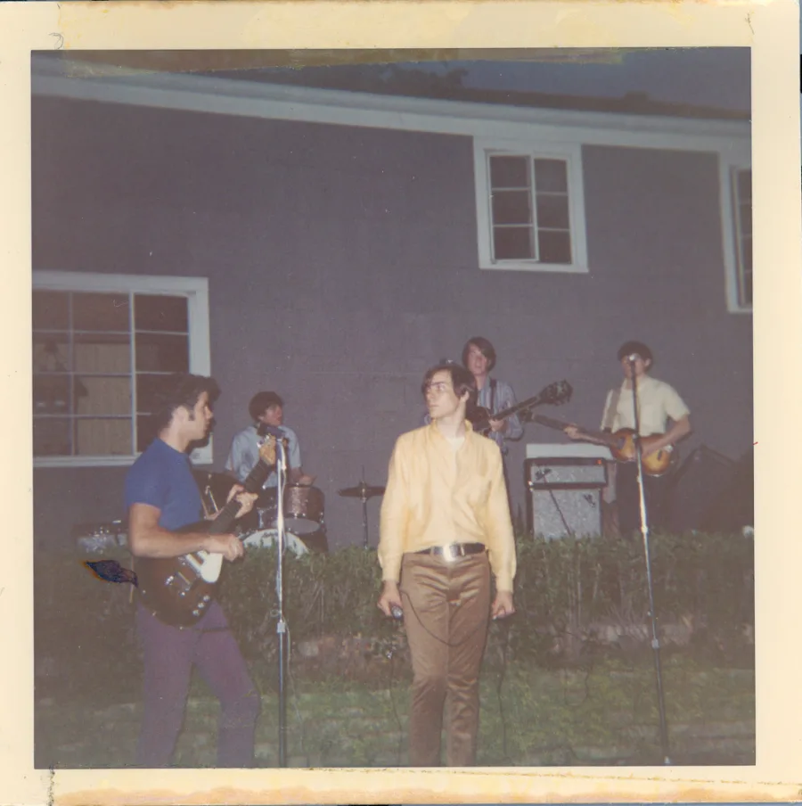 Square color print of a backyard performance at dusk: five members playing in front of a gray clapboard house, guitarist in a blue tee in the foreground, singer in yellow at center, drums and amps along the hedge