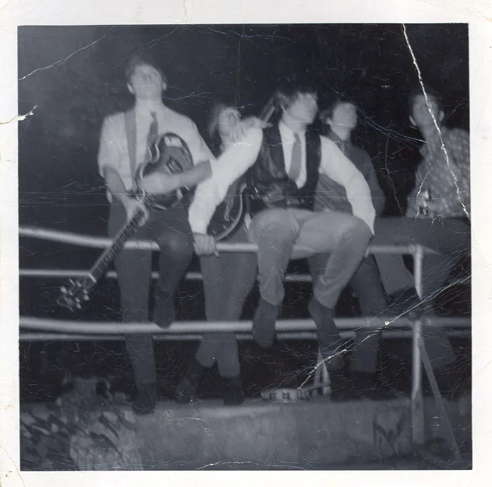 Black-and-white photograph of five boys in ties sitting in a row on a metal railing at night, one cradling a guitar, the print heavily creased