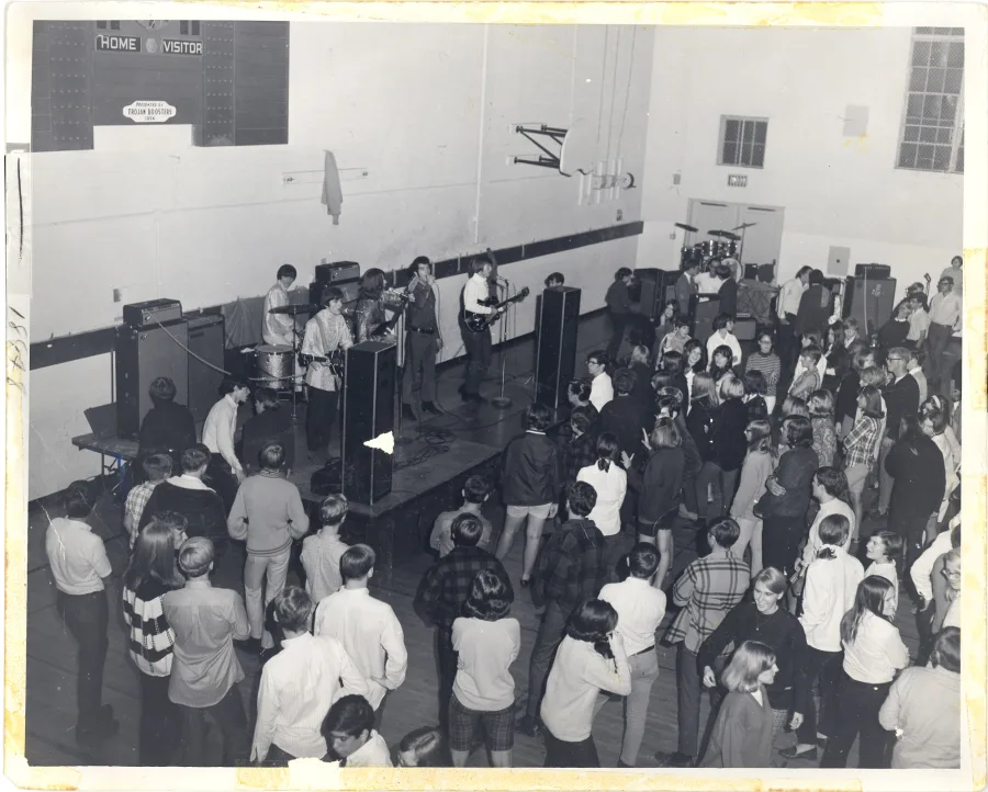 Black-and-white photograph from a balcony of a packed high-school gymnasium dance, HOME and VISITOR scoreboard on the wall, the band playing on a low stage at left over a dense crowd in 1960s dress
