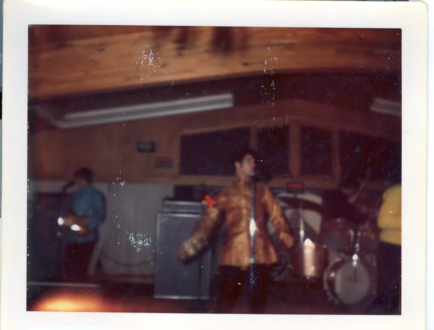 Color print of the singer in the gold brocade jacket mid-move at center stage, arm extended, drum kit at right and a second vocalist at left, under the same wood-paneled lodge ceiling