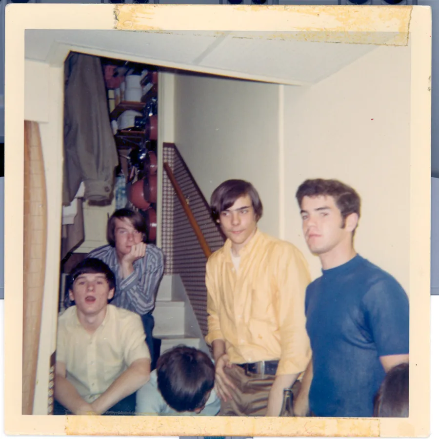 Square color print of five band members crowded on a basement stair landing between sets, in yellow and blue shirts, coats and shelves visible up the staircase behind them