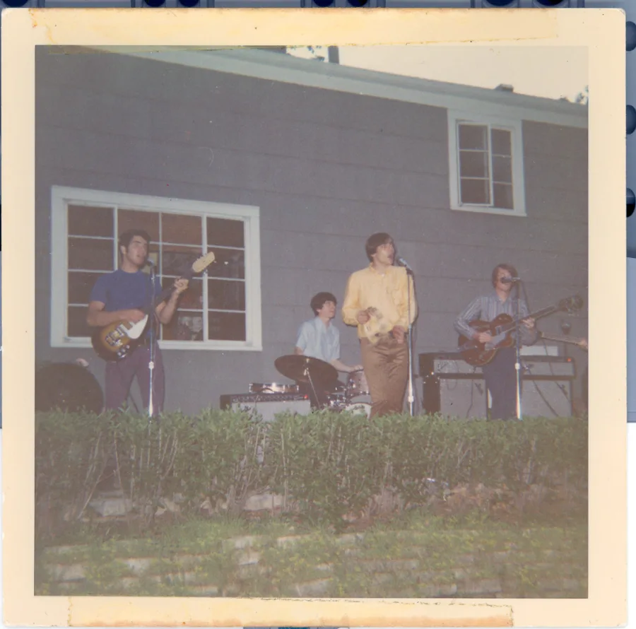 Square color print, same backyard show from further back: the band on a terrace above a stone wall and hedge, guitarist left, drummer center, singer in yellow at the microphone, second guitarist at right