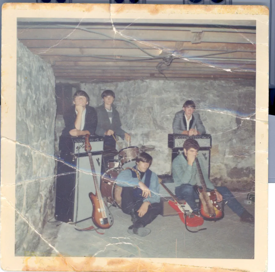Square color print, heavily creased, of the five posed in a stone-walled basement with Fender amplifiers, a drum kit, and two bass guitars laid across the floor