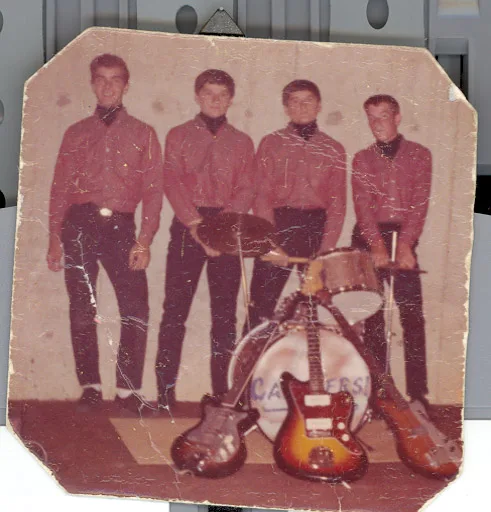 Small color print, corners clipped: four young men in matching dark red shirts and black slacks stand behind a drum kit, three electric guitars propped against it, the drum head hand-lettered with an earlier band name now worn illegible